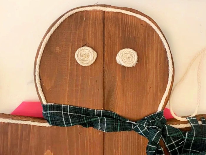 Close-up of a wooden gingerbread man’s face with macrame cord eyes and icing outline, wearing a handmade green tartan scarf.