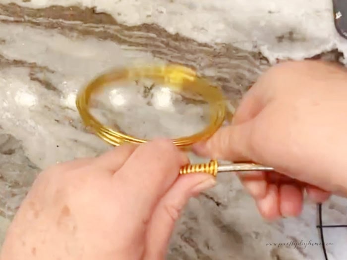 Close-up of gold aluminum wire being wrapped around a metal wreath ring. Leanna’s hands are bending the wire along the top and back side of the ring, with several coils already in place and the remaining wire roll visible in the background.