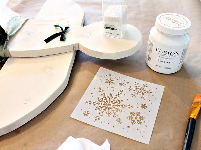 Supplies for decorating the wooden Christmas angel’s wings, including white Fusion paint, a stencil, a paintbrush, and fine iridescent glitter.