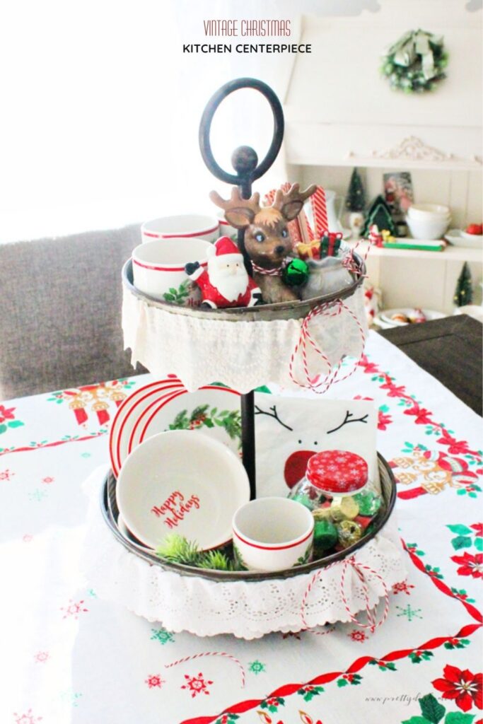 Vintage Christmas centerpiece made with a tiered tray decorated with lace, holiday mugs, small bowls, and festive details on a vintage tablecloth.