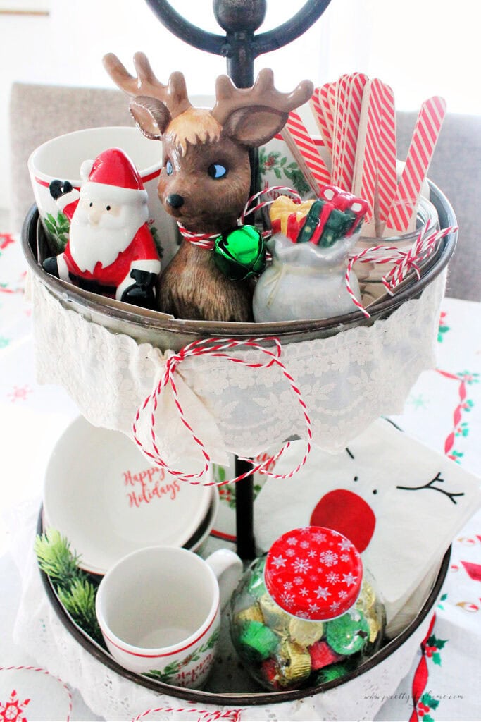 Vintage Christmas tiered tray centerpiece with lace trim, holiday mugs, and small festive dishes arranged on both tiers.