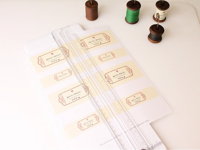 Printed sheet of Santa Thread Company labels being trimmed with a paper cutter, with four wooden spools beside it ready for wrapping.
