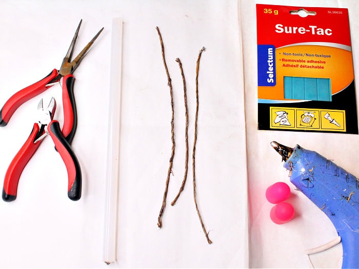 Photo holder craft supplies including needle-nose pliers, wire cutters, paper-covered wire in various lengths, poster putty, a hot glue gun, and silicone finger protectors arranged on a work surface.