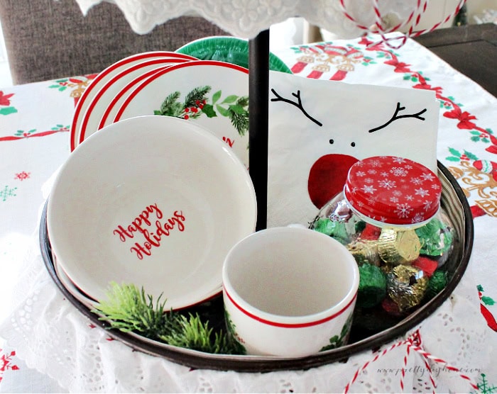 Close-up of the lower tier of a Christmas tray showing vintage-style mugs, a bowl with “Happy Holidays,” wreath plates, cartoon reindeer napkins, and a red-lidded candy jar.