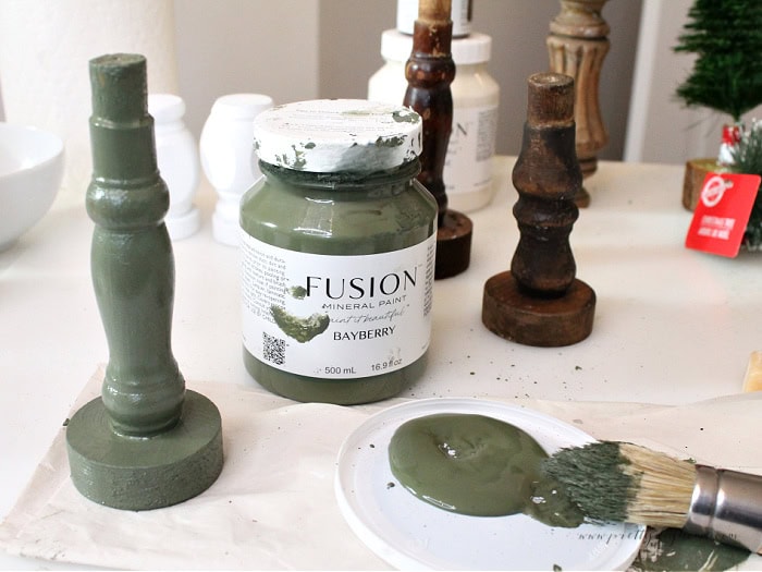 Painting a spindle with Fusion Mineral Paint Bayberry, with other spindles and supplies in the background.