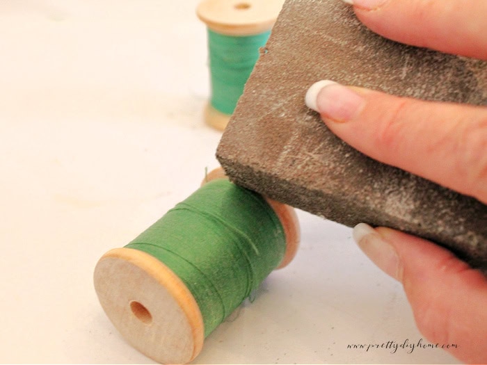 Sanding the edge of a wooden thread spool with a small sanding block to remove old finish and smooth the surface near the remaining thread.