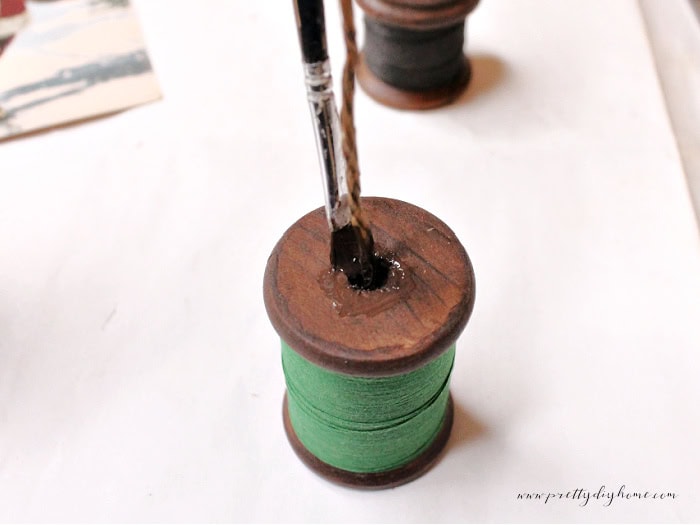 A wooden thread spool with green thread on the base being stained inside using a small brush, with poster putty filling the center to hide the poster putty.