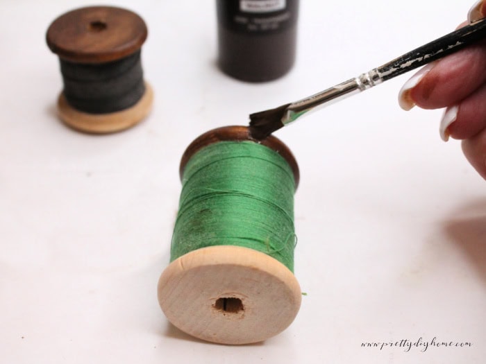 Applying dark walnut stain to the top edge of a wooden thread spool with a small paintbrush, carefully blending the finish near the remaining thread.