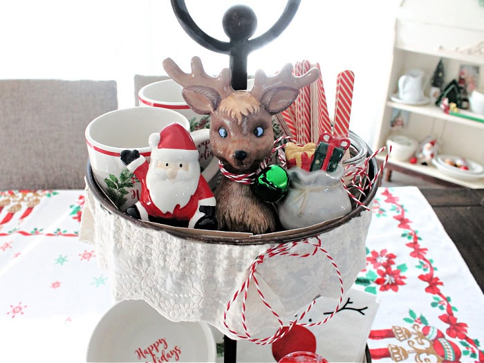 Top tier of a Christmas tray with mugs, a gift bag salt and pepper shaker set, and a mason jar filled with candy cane striped wood cutlery.