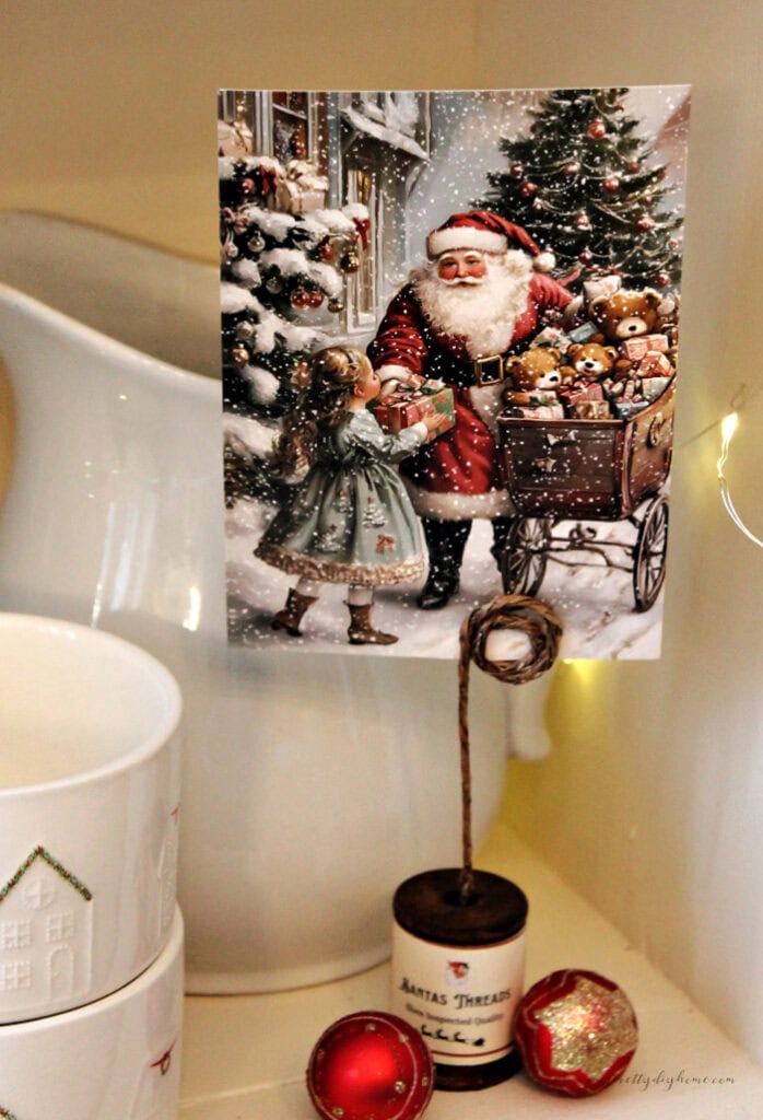 Vintage wooden spool postcard holder with Santa postcard, white ironstone jug, and twinkle lights in a cozy Old-World Christmas display.