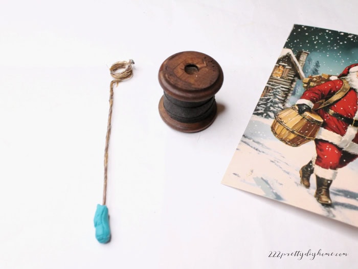 Three parts of the postcard holder craft: a length of paper-covered wire with a curled top and poster putty base, a stained wooden spool, and a vintage Santa postcard ready for assembly.
