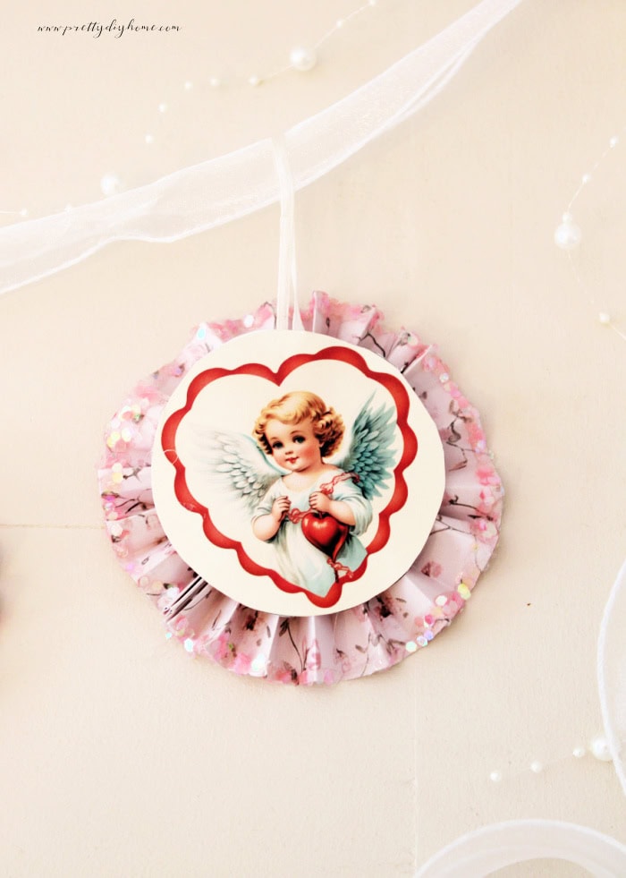 Finished vintage Valentine paper medallion with cupid center, white ribbon hanger, and a string of white pearls styled as a banner