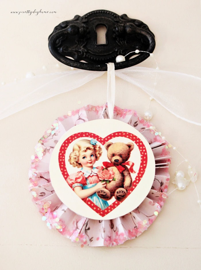 Vintage Valentine paper medallion with a cupid holding a teddy bear, hanging from a metal door pull on a vintage cabinet