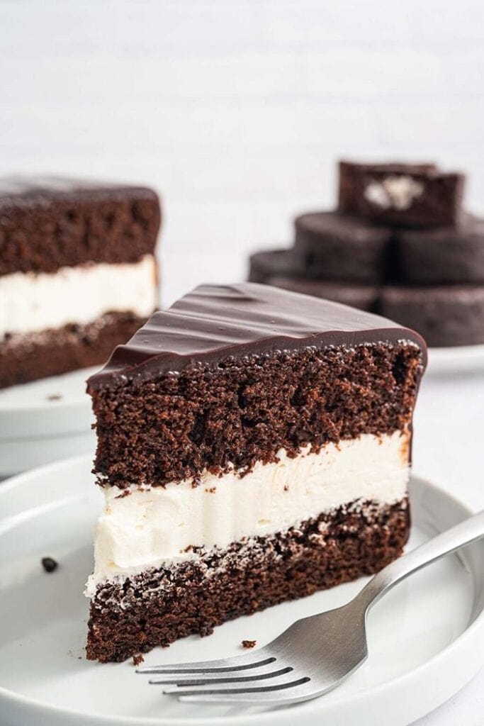 A thick slice of Ding Dong cake with two layers of chocolate cake and a generous fluffy white filling, coated in a thin layer of chocolate ganache, with the whole cake and a pile of chocolate in the background.