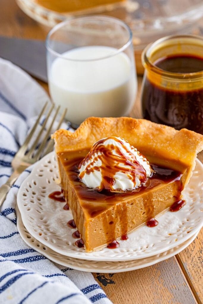 A slice of butterscotch pie sitting on a white plate with a dollop of whipped cream and a drizzle of butterscotch sauce, with a glass of milk and a jar of sauce in the background.
