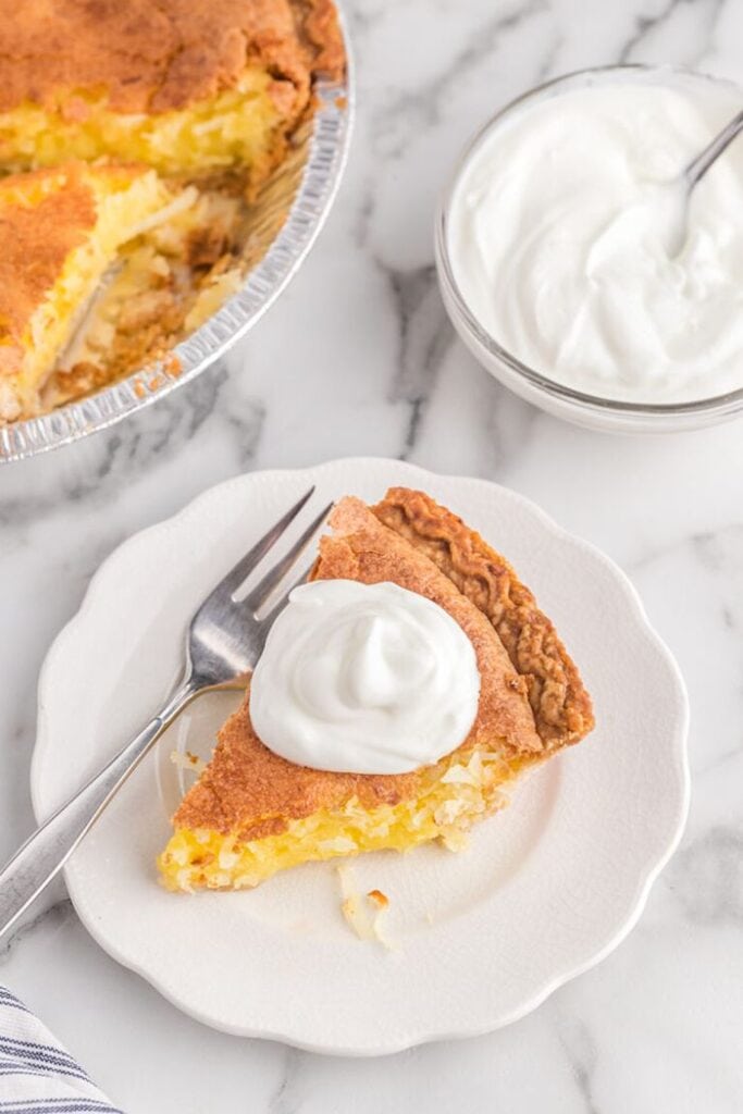A slice of classic coconut pie served on a white plate with a fork, with the whole pie and whipped cream nearby.