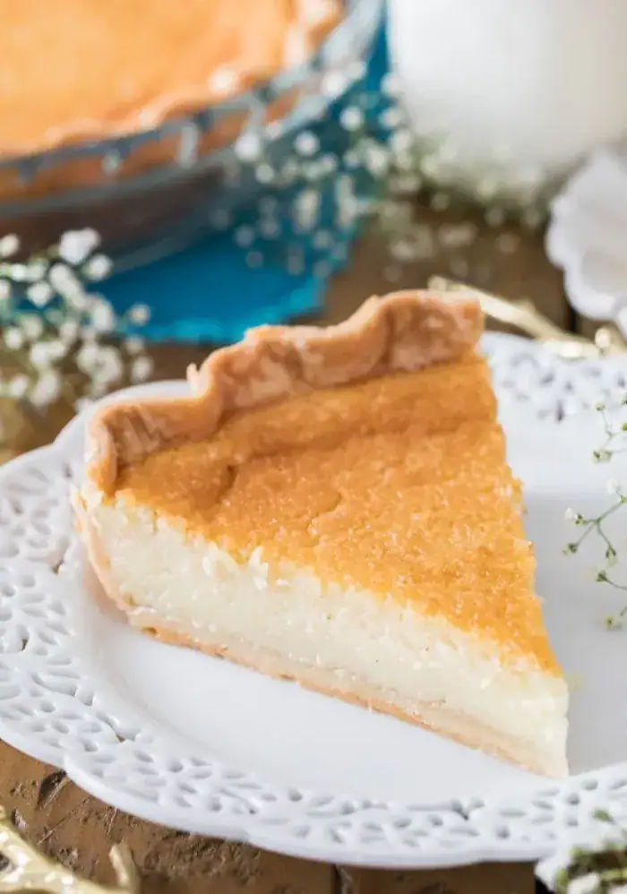 A slice of buttermilk pie with a creamy white filling and golden brown top, with baby’s breath and a blue napkin in the background.