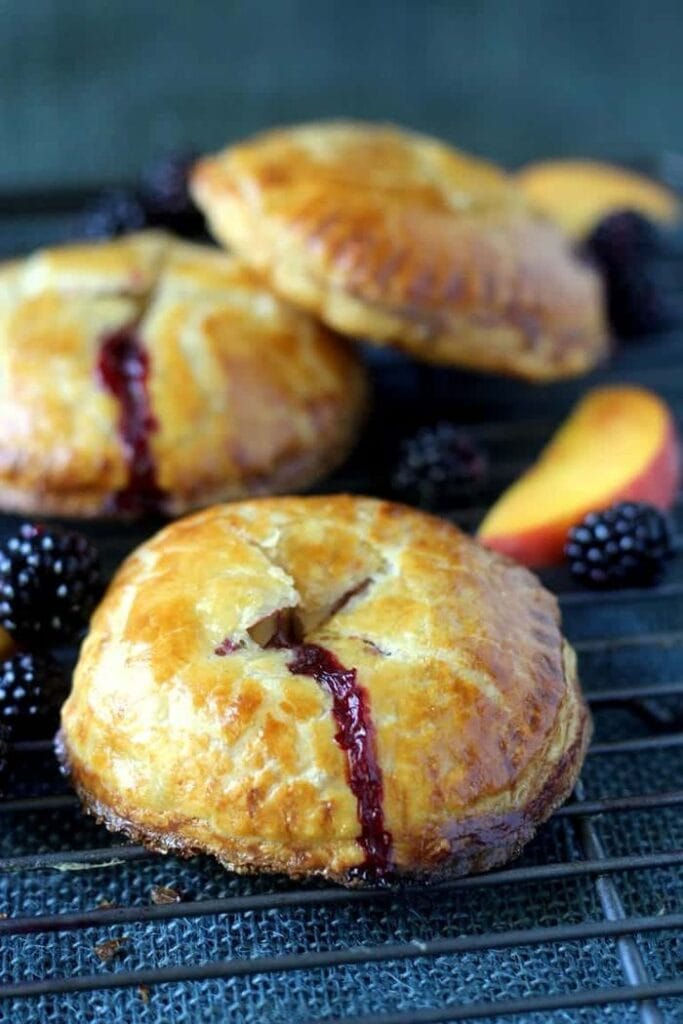 A stack of golden brown round hand pies with blackberry filling oozing from the center, served on a dark towel and garnished with fresh blackberries and peach slices.