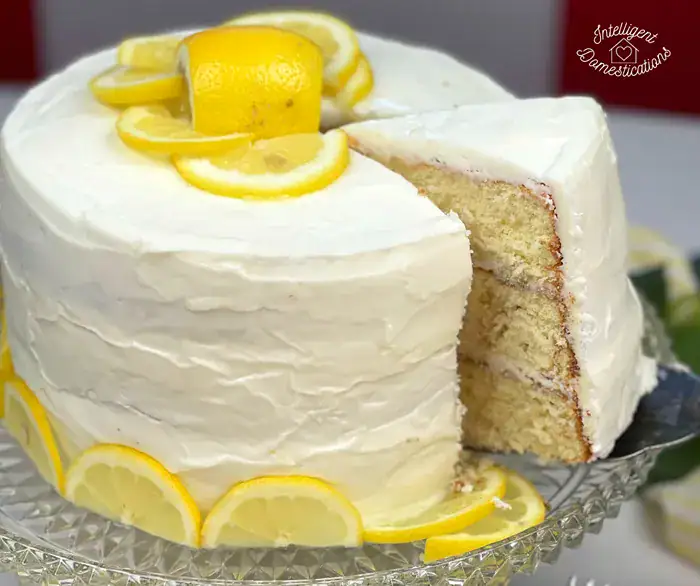 A large three-layer lemon cake decorated with fresh lemon slices, with a piece removed from the center to show the moist cake layers inside.