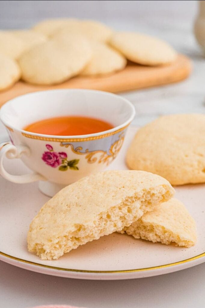 Two old fashioned tea cakes resting on a saucer beside a decorative teacup filled with tea, with one cookie whole and the other broken in half to show the crunchy interior. A wooden tray piled with more tea cakes sits in the background.