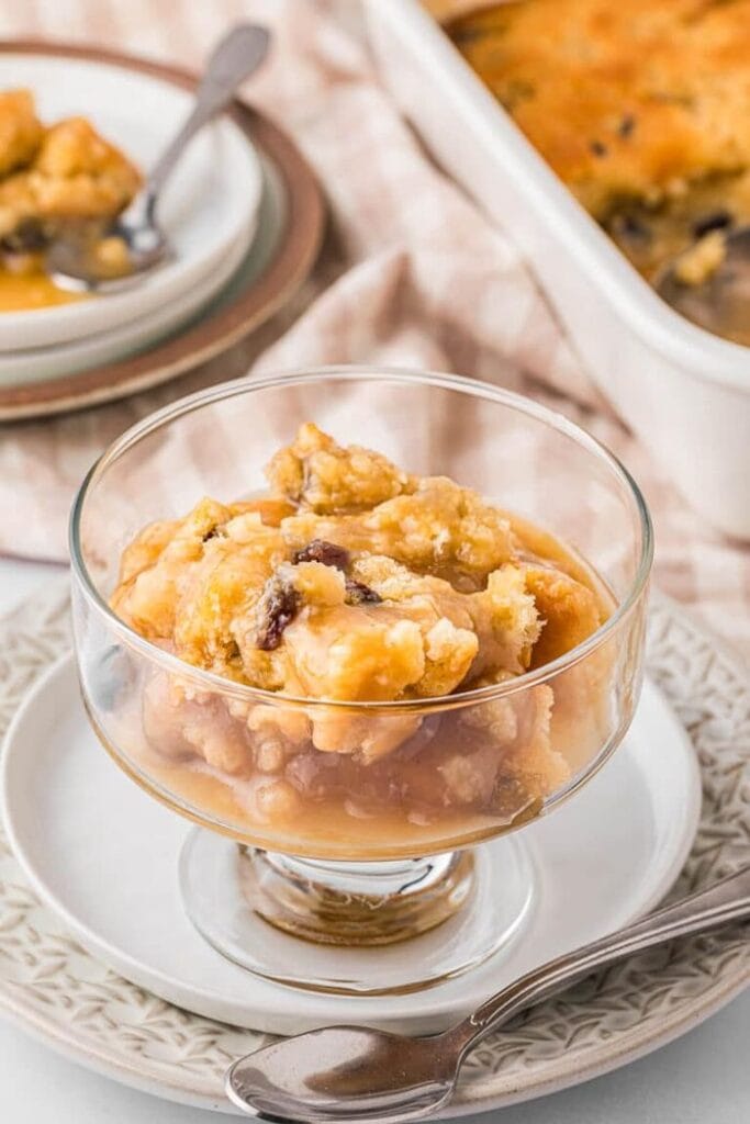 A clear glass dessert dish filled with golden radio pudding, set on a white plate with a tablecloth, with the pudding in a porcelain baking dish and a second serving visible in the background.