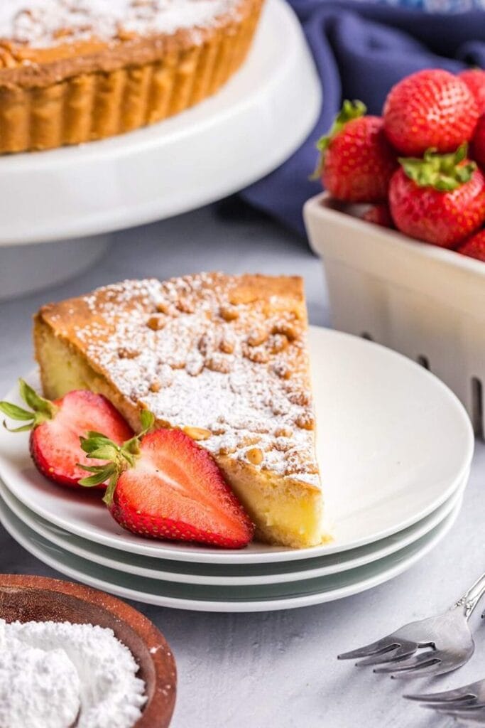 A slice of icing sugar dusted torta della nonna on a stack of three plates, with a halved strawberry beside it, the dessert visible in the background, and a bowl of fresh strawberries nearby.