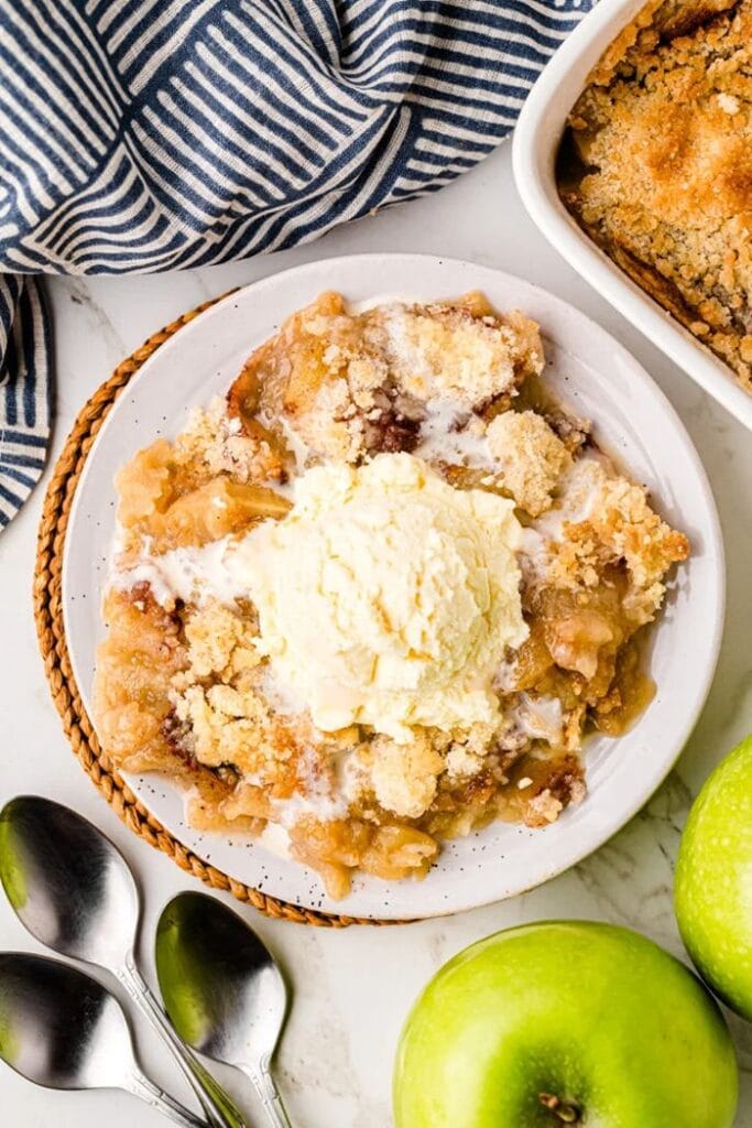A very large serving of apple crisp topped with a scoop of ice cream, shown in an overhead view with a spoon in one corner, a green apple in another, a small portion of the baking pan visible, and a striped napkin alongside.