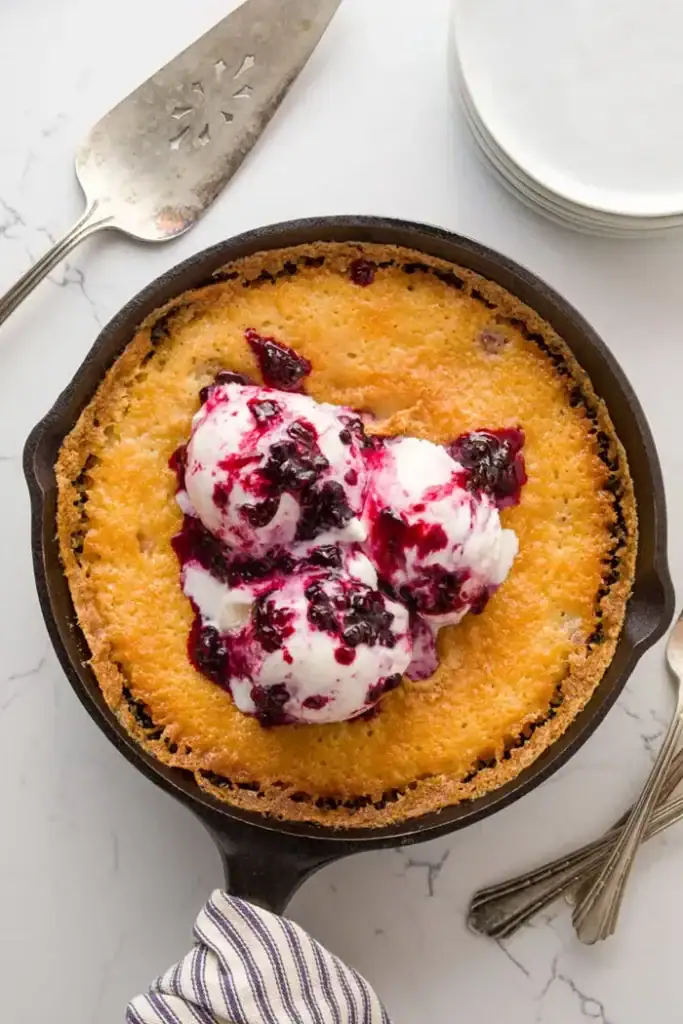 An overhead view of a blackberry cobbler baked in a cast iron pan, topped with whipped cream and fresh blackberries.