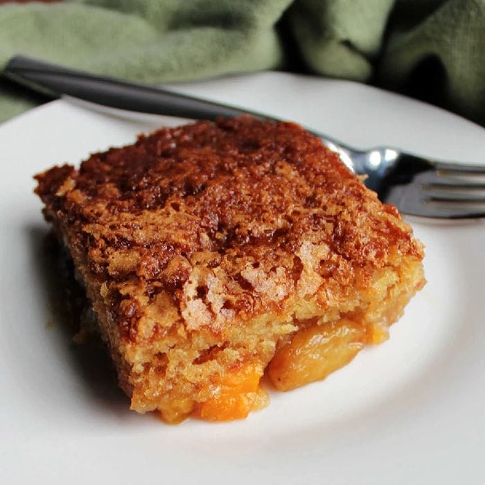 A square slice of fruit cocktail cake served on a white plate with a fork, with a green tea towel in the background.