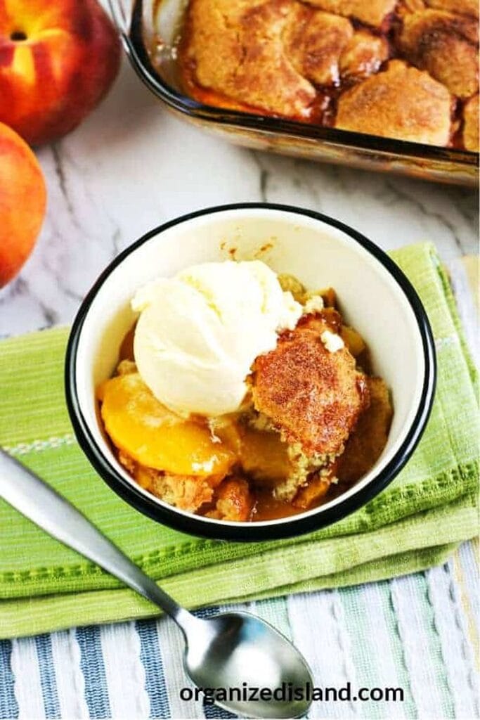 A bowl of peach cobbler with juicy peaches and a golden brown cake topping, served with a scoop of ice cream in a dessert bowl. The bowl sits on a green folded napkin, with fresh peaches and the full pan of cobbler placed alongside.