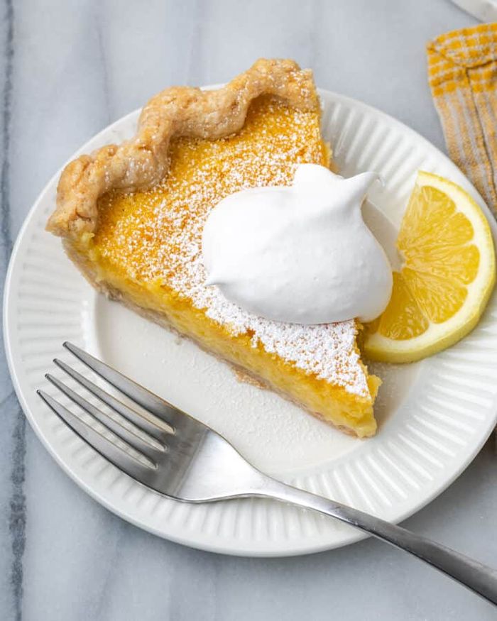 A slightly overhead view of a classic lemon chess pie dusted with icing sugar on a grey marble background, topped with a dollop of whipped cream, a lemon slice on one side of the plate, and a fork resting on the other.