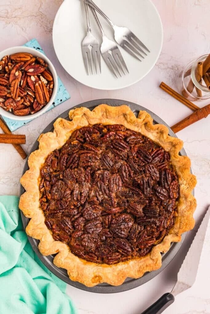 An overhead view of a whole pecan pie, styled with a napkin, a stack of plates with forks, and a bowl of pecans beside it.