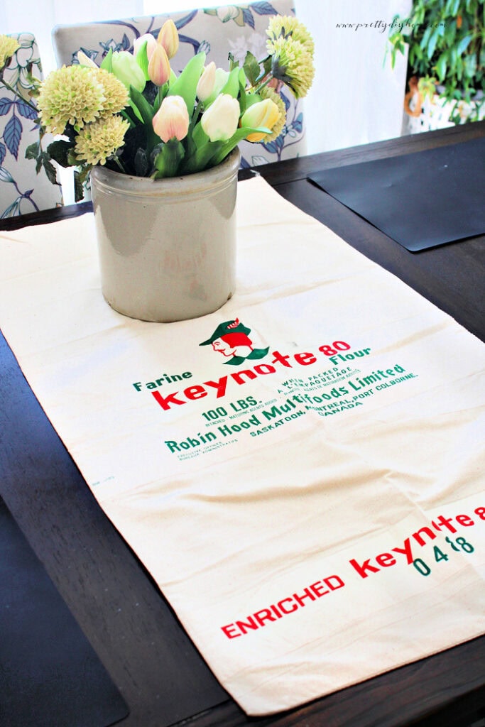 A vintage flour sack used as table runner with crock and flowers.