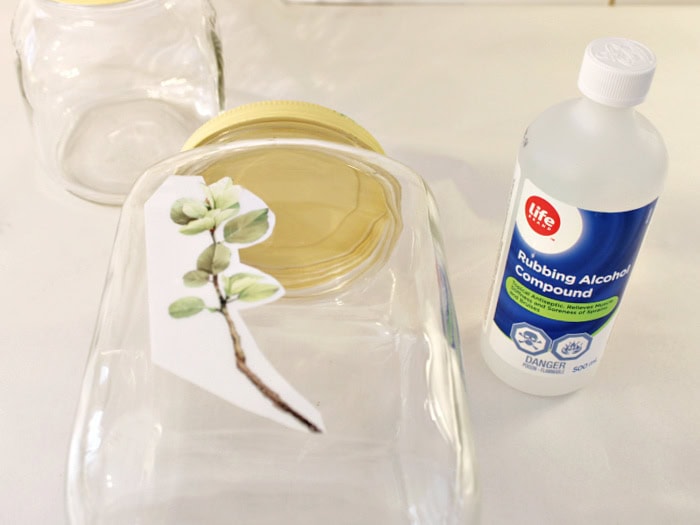 Painted jar with yellow lid and floral transfer placed on glass before applying. A bottle of alcohol used in the jars preparation sitting beside.