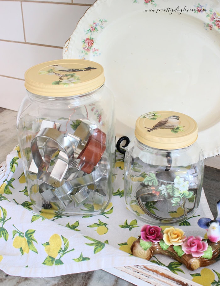 Vintage glass jars with painted lids and bird transfers styled on a cottage kitchen counter.