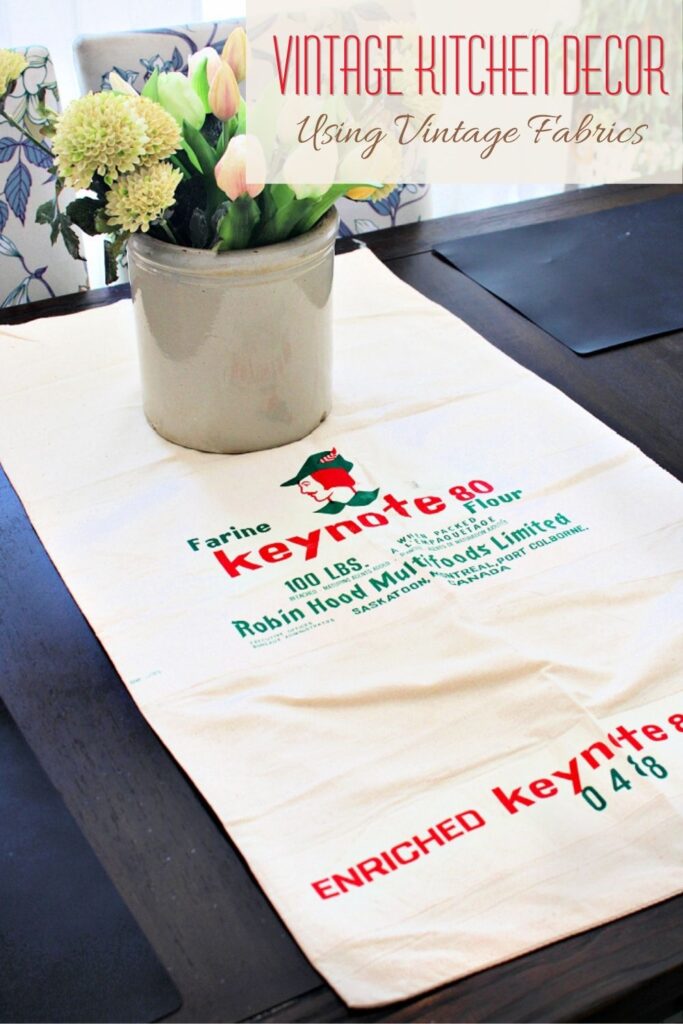 A vintage flour sack used as table runner with crock and flowers in cottage kitchen.