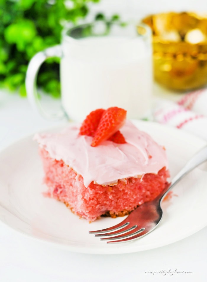 Slice of strawberry slab cake with cream cheese frosting topped with fresh strawberries on a plate with a fork and milk in the background.