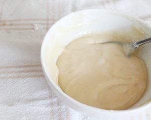 A small white bowl filled with creamy coloured icing for glazing butterhorns. The icing is thin and there is a small teasoon in the icing.