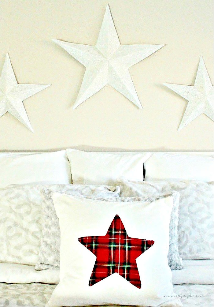 DIY Christmas pillow covers, two are soft grey and the front on is a white cushion cover with red tartan plaid star on the front. All the cushions are lined up on a bed with european cushions in the background and three sparkly stars over the bed.