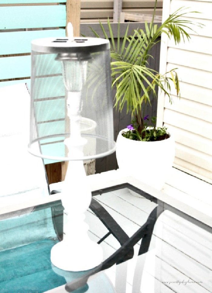 A white outdoor solar lamp with a silver metalic shade. The lamp is sitting on a picnic table with a pretty plant in the back ground.