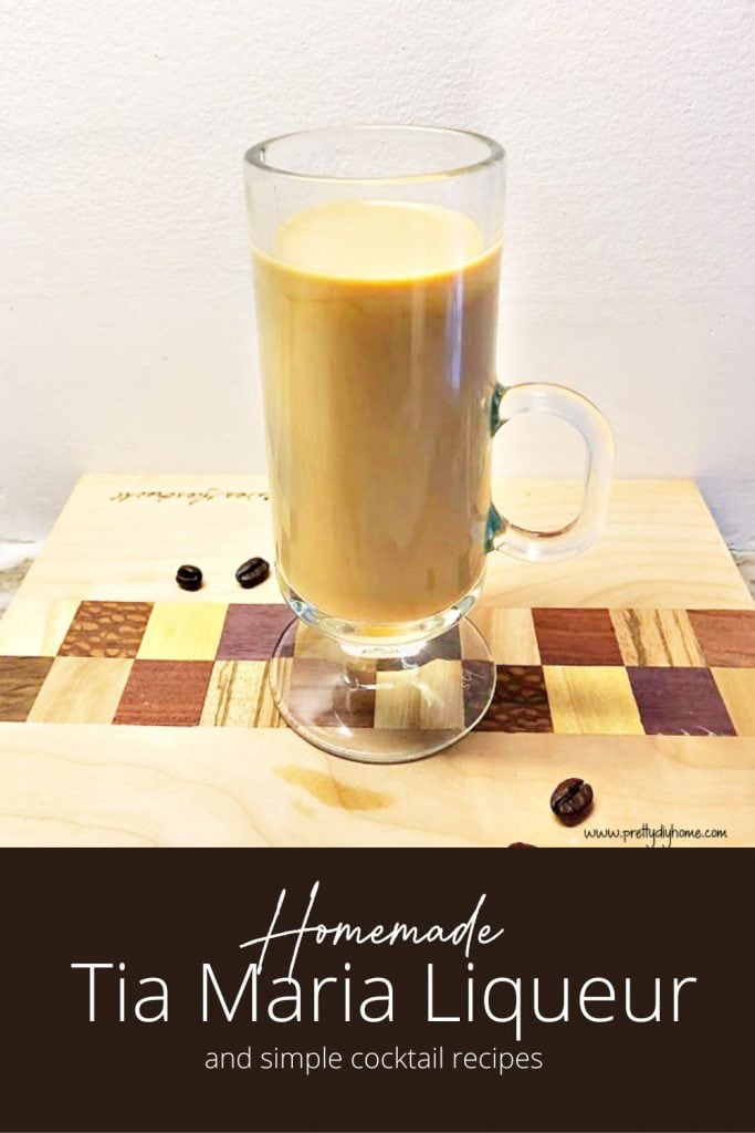A clear tall glass mug filled with tia maria, coffee and cream. The creamy coffee drink is sitting on a wood cutting board with many different types of wood, and the board is scattered with coffee beans.