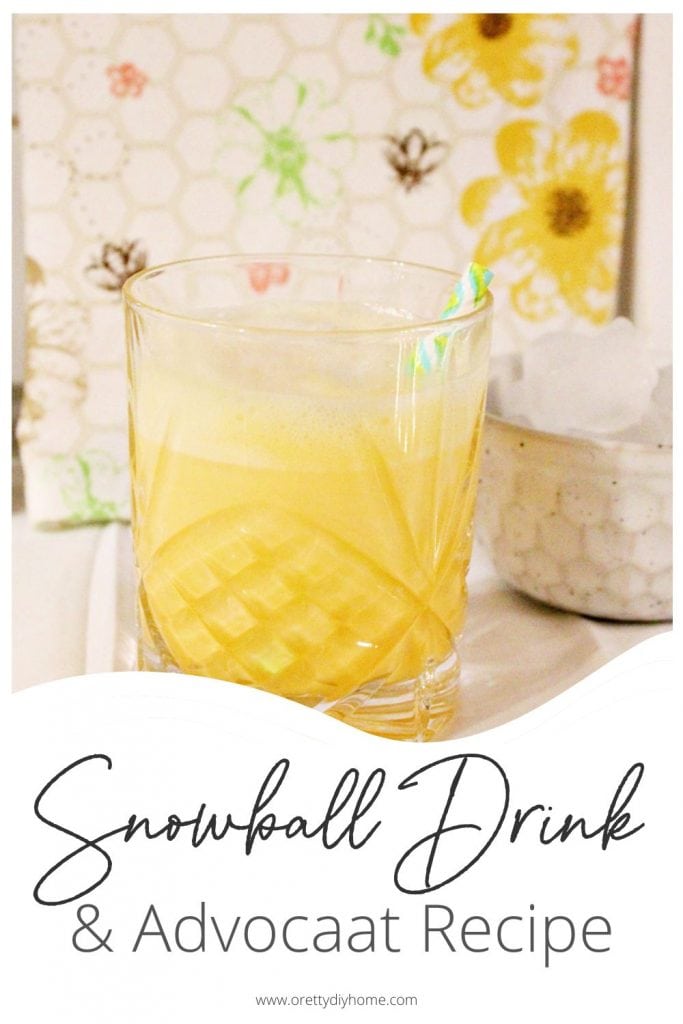 A large crystal cocktail glass filled with bright yellow snowball cocktail made using homemade advocaat. the background is bright colours of pink and yellow flowers and there is a small bowl of ice beside it.