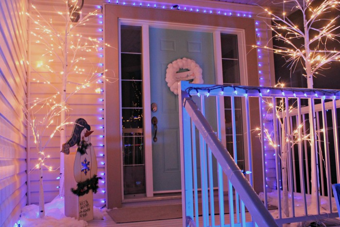 Front porch Christmas lights in blue and white, with blue Christmas light strings around the door and bright white Christmas trees. There is a litttle DIY snowman yard decorations flanking a Christmas door decorated with a DIY snowman wreath.