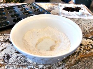 A large miing bowl filled with dry ingredients. There is a well in the middle filled with milk and yeast. You can see currants and raisins in the background along with gugelhupf cake pans.