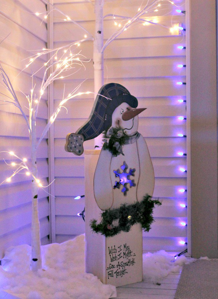 Homemade DIY wooden outdoor Christmas decorations at night. A DIY snowman yard decoration made from wood as an exterior Christmas decorating idea. The little snowman is lit up with pretty blue and white front porch Christmas lights.