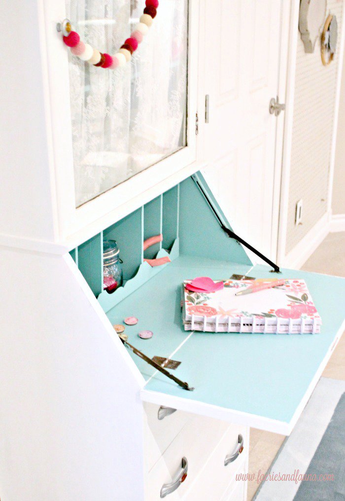 A finished furniture refinishing project in white paint. The interior of the old secretary is painted a soft blue, and it shows the inside cubicles with pretty pink accessories.