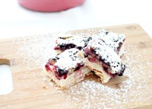 Four lemon blueberry shortbread bars on a wood board. The blueberry lemon bars have a shortbread crust, a fresh blueberry and lemon custard layer and topped with pretty white icing sugar.
