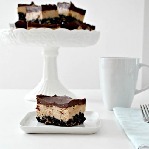 A large tray of No Bake Chocolate Peanut Butter bars with three layers. Oreo layer, peanut butter layer in the center and topped with melted chocolate chips. You can see several cut bars on a tray, one served on a plate with a cup of coffee beside it.