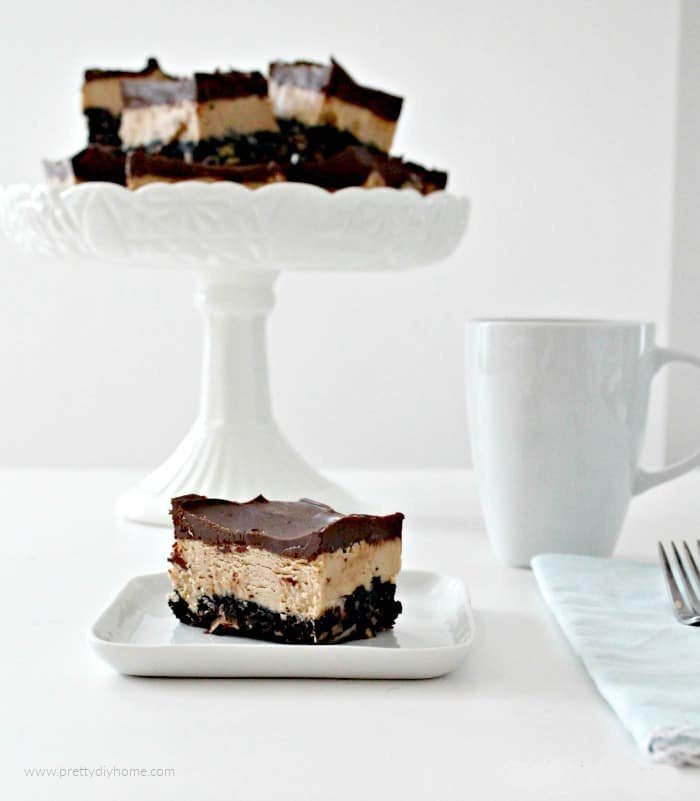 A large tray of No Bake Chocolate Peanut Butter bars with three layers. Oreo layer, peanut butter layer in the center and topped with melted chocolate chips. You can see several cut bars on a tray, one served on a plate with a cup of coffee beside it.