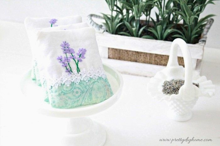 Lavender sachets displayed on a white pedestal plate with lavender and a milk glass basket in the background.